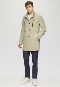 Lichtgroene jas met hoge kraag, drie zwarte knopen, een ritszak, gecombineerd met donkere broek en witte sneakers. Gladde textuur.