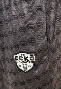 Svart stoff med grå sirkulære mønstre har et merkelapp med "ECKO UNLIMITED" og et stilisert grafisk design. Myk tekstur, lett design.