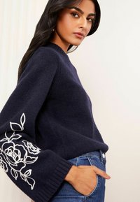 Pulover bleumarin dintr-un material texturat, cu un corp ajustat și mâneci supradimensionate, decorate cu broderii albe cu flori la manșete. Blugi vizibili la talie.