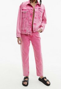 Chaqueta de mezclilla rosa con pantalones de mezclilla rosa a juego, con un corte relajado y bolsillos frontales. Sandalias negras completan el atuendo.