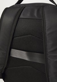 Le matériau du sac à dos noir présente un maillage texturé et un tissu lisse, avec une sangle grise horizontale et des motifs géométriques sur le panneau arrière.
