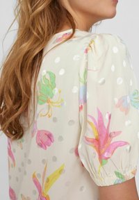 Kvinna med långt vågigt hår som bär en krämfärgad blus med puffärmar, silverfärgade prickar och flerfärgade blommönster.