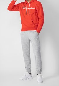 Czerwona bluza z kapturem z białym logo "Champion", w komplecie z szarymi dresami z elastycznymi mankietami i białymi sneakersami. Prosty, casualowy design.