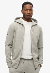 Grå zip-hoodie i blødt stof med en frontlomme og snøre i hætten, parret med en hvid skjorte og matchende grå bukser.