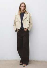 Beige beskuren dubbelknäppt jacka med vida ärmar, kombinerad med mörkbruna vida byxor. Platta svarta sandaler fulländar looken.