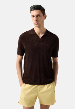 Man met een donkerbruin poloshirt met korte mouwen, lichtgele korte broek en zwarte zonnebril op zijn hoofd, staand met handen in de zakken.