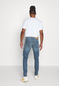 Jean bleu skinny avec surpiqûres contrastées, étiquette à la taille, porté avec un t-shirt blanc uni et des baskets de couleur claire.