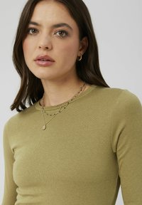 Haut à manches longues vert olive avec une texture lisse ; colliers en or superposés avec un petit pendentif et des perles noires. Encolure ronde subtile.