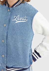 Džínová bomber bunda s bílými rukávy, námořnickým ribovaným límcem a manžetami. Obsahuje vyšitý logo Levi's na hrudi a patentky.