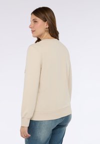 Béžová mikina s dlouhými rukávy, jemnou texturou a žebrovanými manžetami. Zadní pohled ukazuje kulatý výstřih a volný střih v kombinaci s modrými džíny.