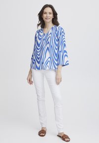 Bluză albă cu mâneci lungi, cu un model unduit în nuanțe de albastru și alb; material ușor; combinată cu pantaloni albi și sandale plate maro.