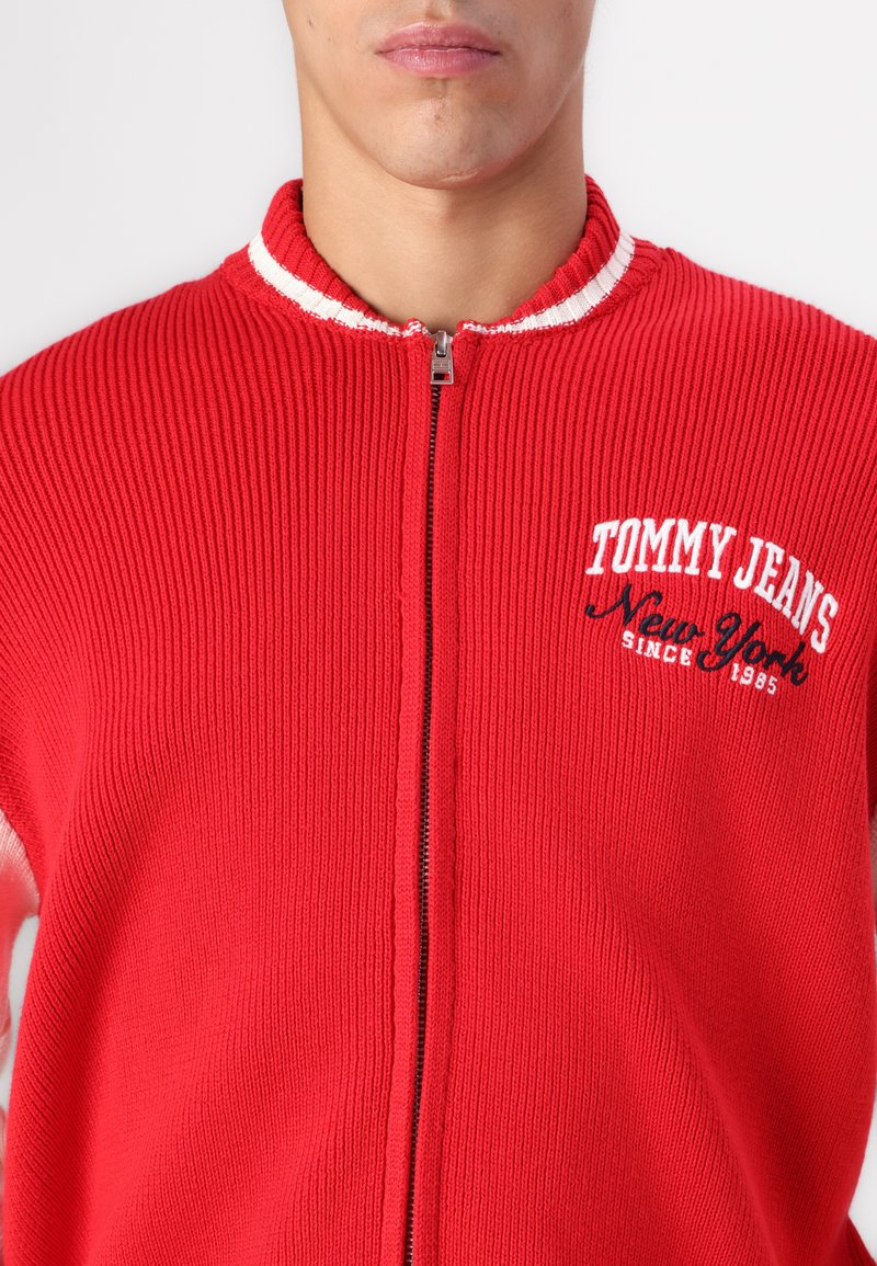 Rote gerippte Zip-Jacke mit weißem gestreiften Kragen. Auf der Vorderseite mit "TOMMY JEANS New York seit 1985" in Weiß und Schwarz bestickt.