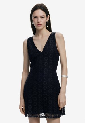 Spitzenkleid in tiefem Marineblau, mit V-Ausschnitt, ärmellosem Design, floralem Muster und einer figurbetonten Silhouette, akzentuiert durch ein silbernes Armband.