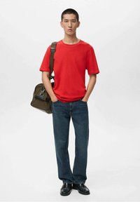 Červené bavlnené tričko, vo voľnom strihu, skombinované s tmavomodrými džínsami. Model má na sebe olivovozelenú tašku cez plece a čierne topánky. Jednoduchý dizajn.