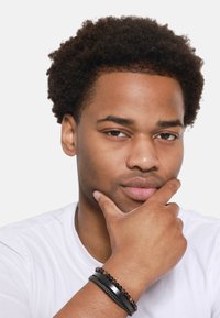 Des bracelets noirs et un accent en métal ornent une main reposant sous un menton ; un T-shirt blanc contraste avec des cheveux bouclés foncés et un teint de peau.