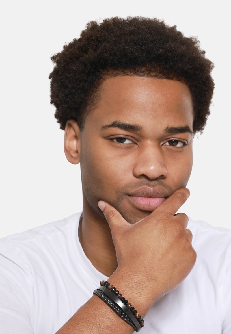 Des bracelets noirs et un accent en métal ornent une main reposant sous un menton ; un T-shirt blanc contraste avec des cheveux bouclés foncés et un teint de peau.
