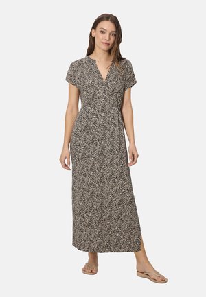 Mujer de pie con vestido largo de manga corta, estampado en blanco y negro, con cuello en V y sandalias beige de dedo, sobre un fondo claro.