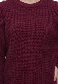 Gros plan d'une personne portant un pull en maille texturé rouge foncé avec un col rond et des manches longues.
