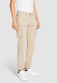 Beige Cargo-Hosen mit elastischen Knöchelbündchen, mehreren Seitentaschen und einem Knopfverschluss, hergestellt aus strapazierfähigem Stoff. Kombiniert mit weißen Sneakers.