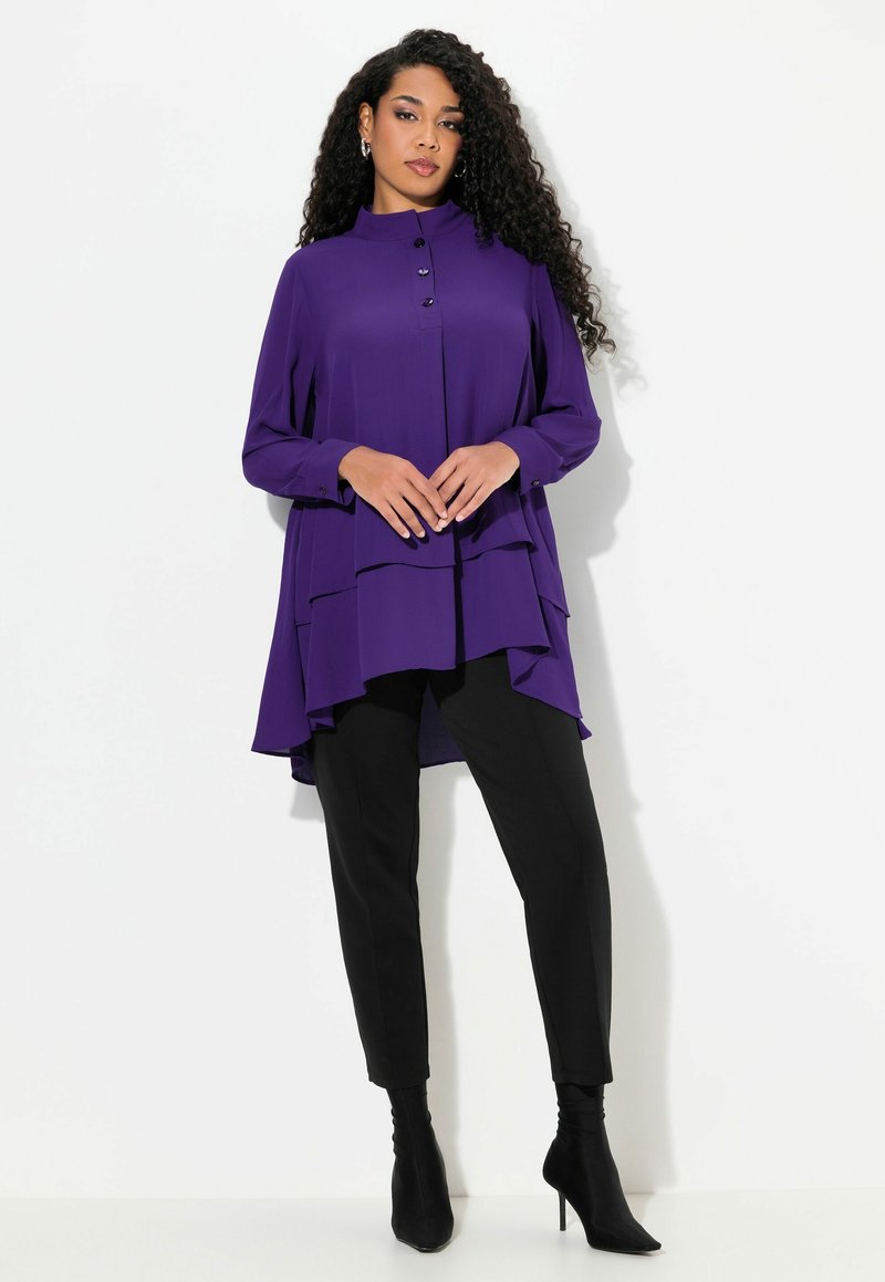 Fialová vrstvená tunika s vysokým límcem a knoflíky, spárovaná s černými úzkými kalhotami a černými lodičkami ke kotníkům, na bílém pozadí.