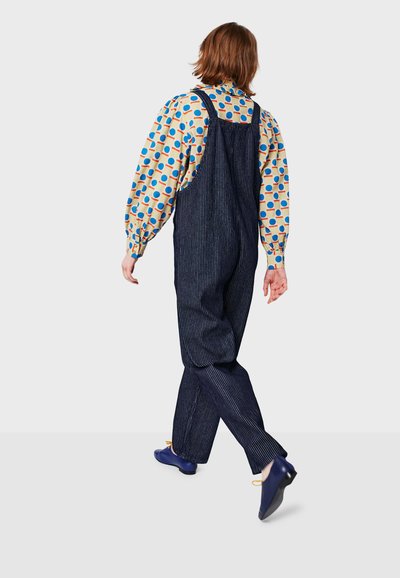 Dark blue pinstripe overalls with adjustable straps, worn over a light-colored shirt featuring colorful polka dots. Blue flat shoes.