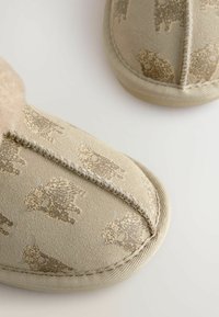 Chaussons beige avec une surface douce et texturée, présentant un subtil motif animal en brun. Finitions avec des coutures détaillées et un bout arrondi.