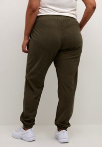 Pantaloni jogger verde oliva con vita elastica e polsini alla caviglia, realizzati in tessuto morbido. Sneakers bianche visibili nella parte inferiore.