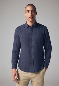 Camisa de botones de manga larga, azul oscuro, con tela texturizada, bolsillo en el pecho y botones marrones; combinada con pantalones caquis.
