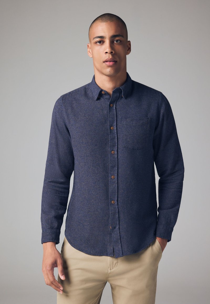 Camisa de botones de manga larga, azul oscuro, con tela texturizada, bolsillo en el pecho y botones marrones; combinada con pantalones caquis.