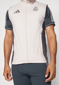 Chaleco acolchado rosa claro con detalles en gris, cierre de cremallera y cuello alto. Presenta el logotipo del equipo en el pecho y diseño de tres rayas en los hombros.