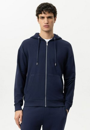 Junger Mann trägt einen marineblauen Kapuzenpullover mit Reißverschluss und Fronttaschen sowie passende marineblaue Hosen vor einem einfarbigen hellen Hintergrund.