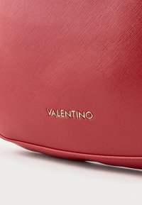 Crvena kožna torba s teksturiranom površinom, koja sadrži zlatni "Valentino" logo u sofisticiranom fontu, s glatkom završnom obradom i zaobljenim rubovima.