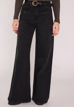 Femme portant un jean noir taille haute à jambes larges avec des poches à rabat à l'avant et une ceinture noire, associé à un haut marron à manches longues.