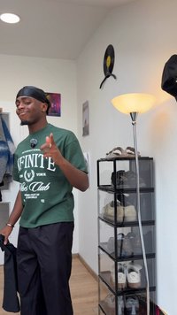 Jovem em camisete gráfica verde e calças pretas aponta o dedo dentro de um quarto com armazenamento de sapatos e uma lâmpada de pé.