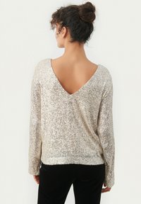 Schimmernder silberner Pullover mit tiefem V-Ausschnitt hinten, langen Ärmeln und Struktur aus Pailletten. Weicher Stoff, lässige Passform und gerippte Saumdetails.