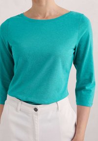 Turkos bomullströja med rundad halsringning och trekvartslånga ärmar, tillsammans med vita högmidjade byxor med knappdetalj.