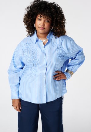 Femme aux cheveux bouclés portant un chemisier bleu rayé avec broderie florale et un pantalon bleu marine, debout, une main sur la hanche.