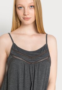 Top cinza sem mangas com alças finas, apresentando um detalhe de renda em padrão zig-zag na gola e um tecido plissado para um ajuste solto.