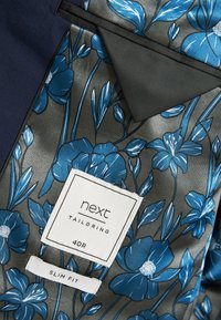 Maßgeschneiderte Jacke von innen mit floralem Muster in Blau auf grauem Hintergrund. Etiketten mit "next", "40R" und "Slim Fit". Texturierte Oberfläche.