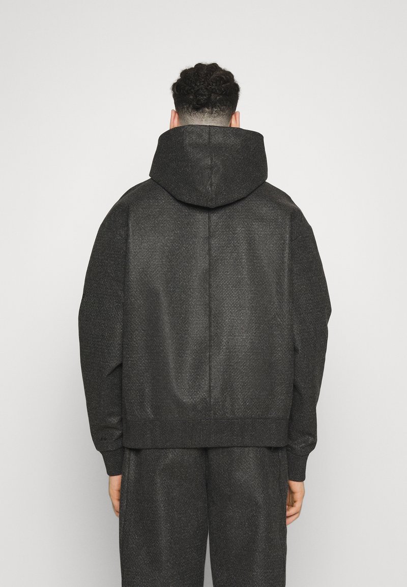 Chaqueta negra con capucha, de tejido texturizado, corte holgado y bajo de canalé. Presenta un diseño limpio y detalles minimalistas.