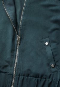 Veste vert foncé avec fermeture éclair sur le devant, texture de tissu lisse et poche latérale avec bouton-pression en argent.