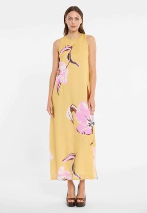 Femme portant une robe jaune sans manches, longue jusqu'au sol, ornée de grandes impressions florales roses, debout les mains détendues le long du corps.