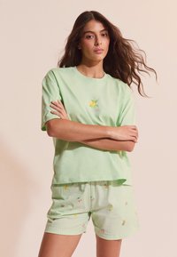 Mujer con cabello largo y ondulado, vestida con una camiseta verde claro con un gráfico de limón y pantalones cortos a juego con pequeños patrones de frutas, de pie con los brazos cruzados.