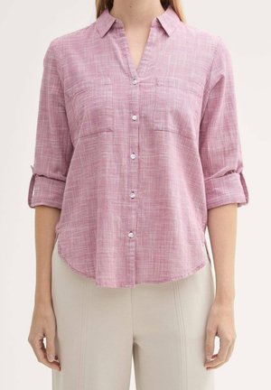 Mujer con camisa rosa de botones con estampado y mangas remangadas y pantalones beige claros, de pie frente a un fondo liso.