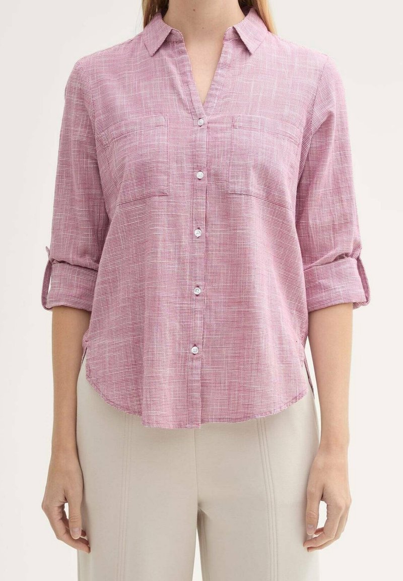 Femme portant une chemise rose à motifs boutonnée avec manches retroussées et pantalon beige clair, debout devant un fond uni.