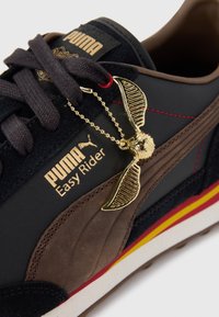 Baskets PUMA Easy Rider avec une tige noire et marron, dotées d'accents dorés, de superpositions texturées et d'un charme décoratif en forme d'aile sur les lacets.