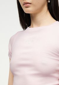 Νεαρή γυναίκα που φοράει ένα ανοιχτό ροζ κοντομάνικο ριμπ τοπ με διακριτικό κέντημα "GUESS JEANS" στο στήθος.