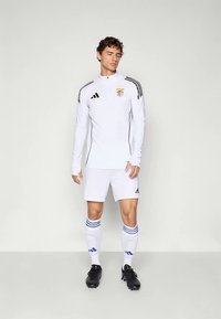 Balti Adidas treniruočių marškinėliai ilgomis rankovėmis, su juodais ir mėlynais akcentais, derinami su baltais šortais ir kelių ilgio kojinėmis su mėlynomis juostelėmis.