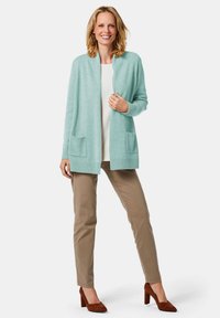 Helle mintgrüne Strickjacke mit langen Ärmeln, zwei Fronttaschen, gerippten Bündchen und Saum, getragen über einem weißen Oberteil und beigen Hosen.