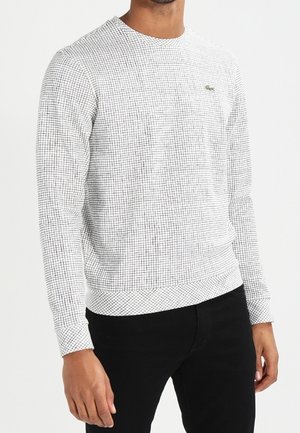 Man draagt een wit overhemd met lange mouwen met klein zwart ruitpatroon en zwarte broek tegen een effen achtergrond.